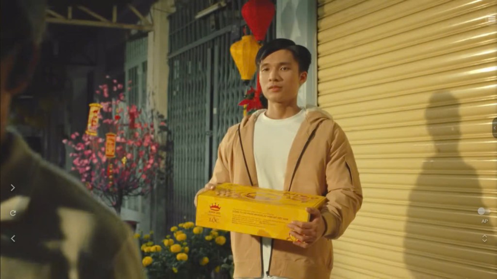 Sự kết hợp giữa tông màu vàng, đỏ trên hộp bánh, trên những bông hoa và chiếc đèn lông cùng màu be trên khung cửa, bộ quần áo của chàng trai, ông chủ tiệm vừa mang đến niềm vui, niềm hy vọng cho năm mới nhưng cũng đầy nét hoài cổ. Ảnh chụp màn hình.